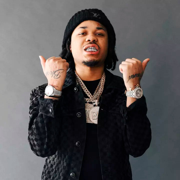 Man in a black beanie and jacket with chains and watches, both thumbs pointing up